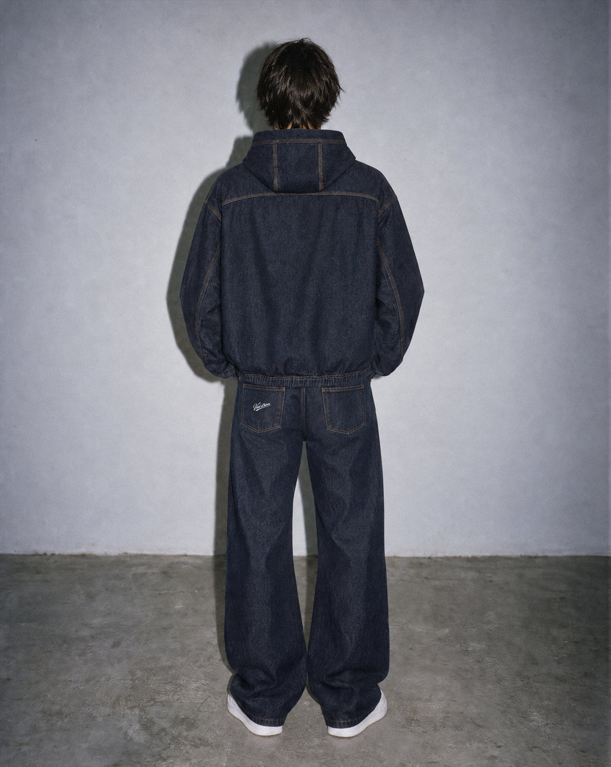 A person viewed from behind wearing a dark indigo denim hooded jacket and matching wide-leg denim pants, standing against a textured gray wall and concrete floor.