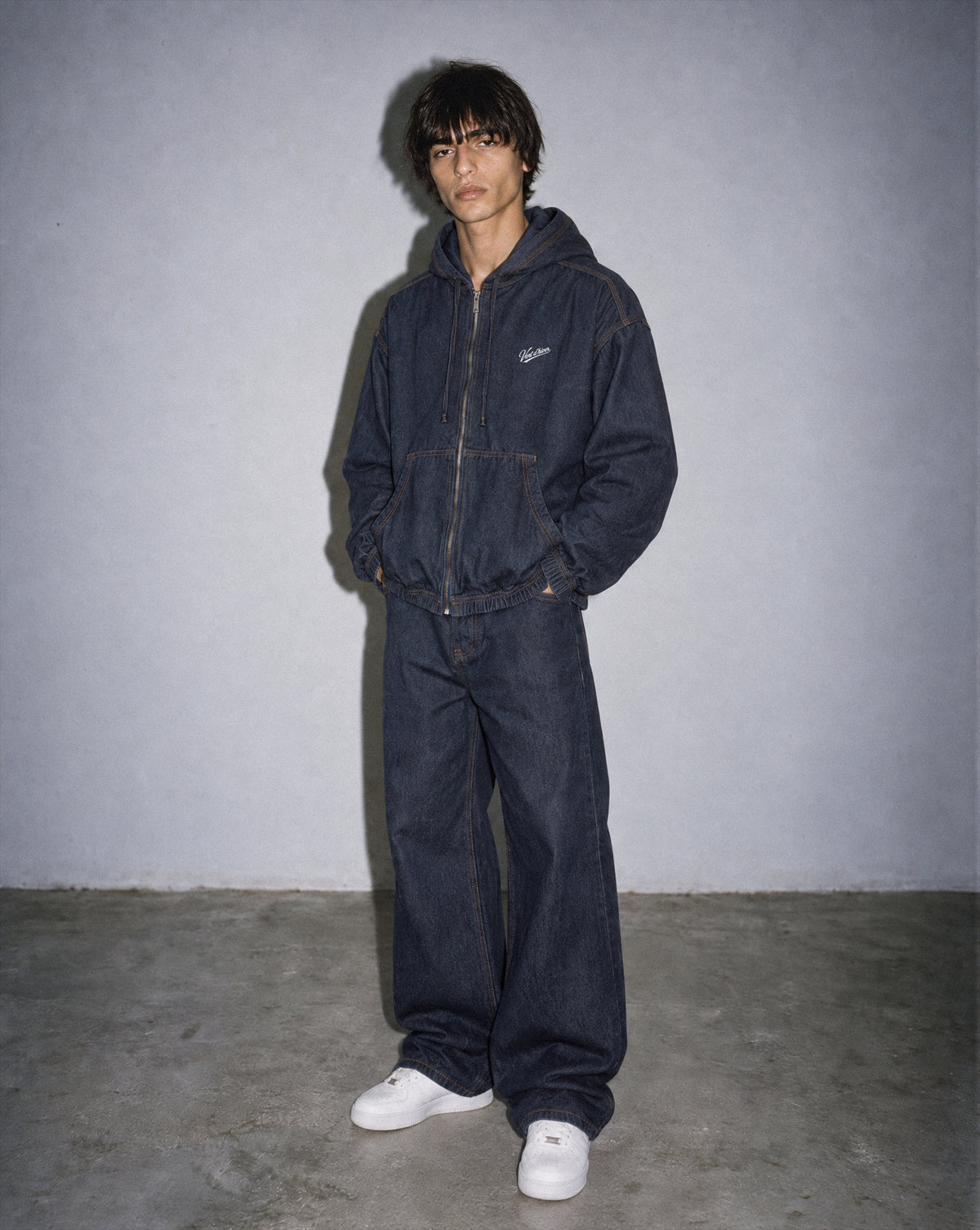 A model stands against a textured gray wall wearing a dark indigo denim hooded jacket and matching wide-leg denim pants, paired with white sneakers.