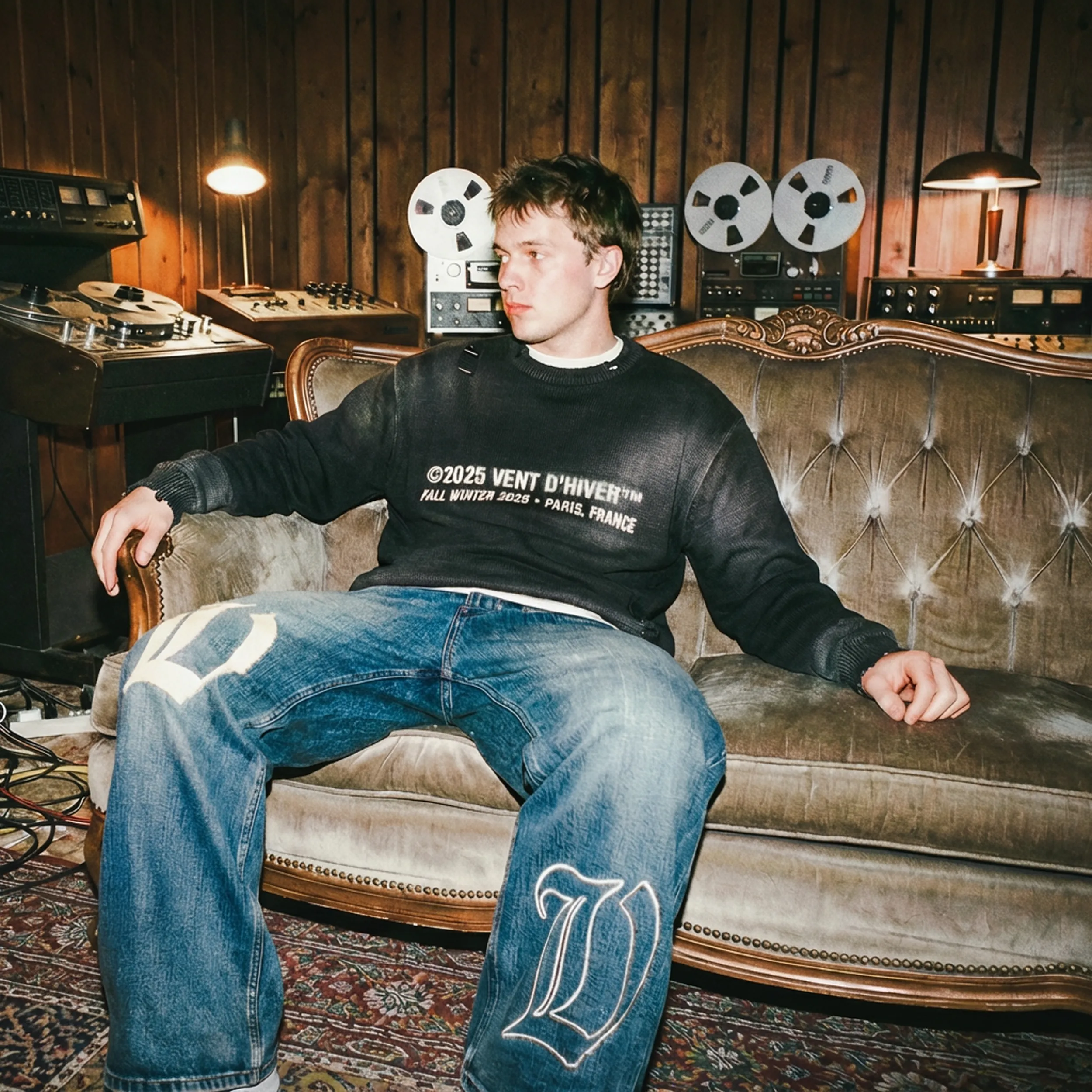 Young man seating on a sofa in a recording studio wearing sweater and jeans from VENT D'HIVER.