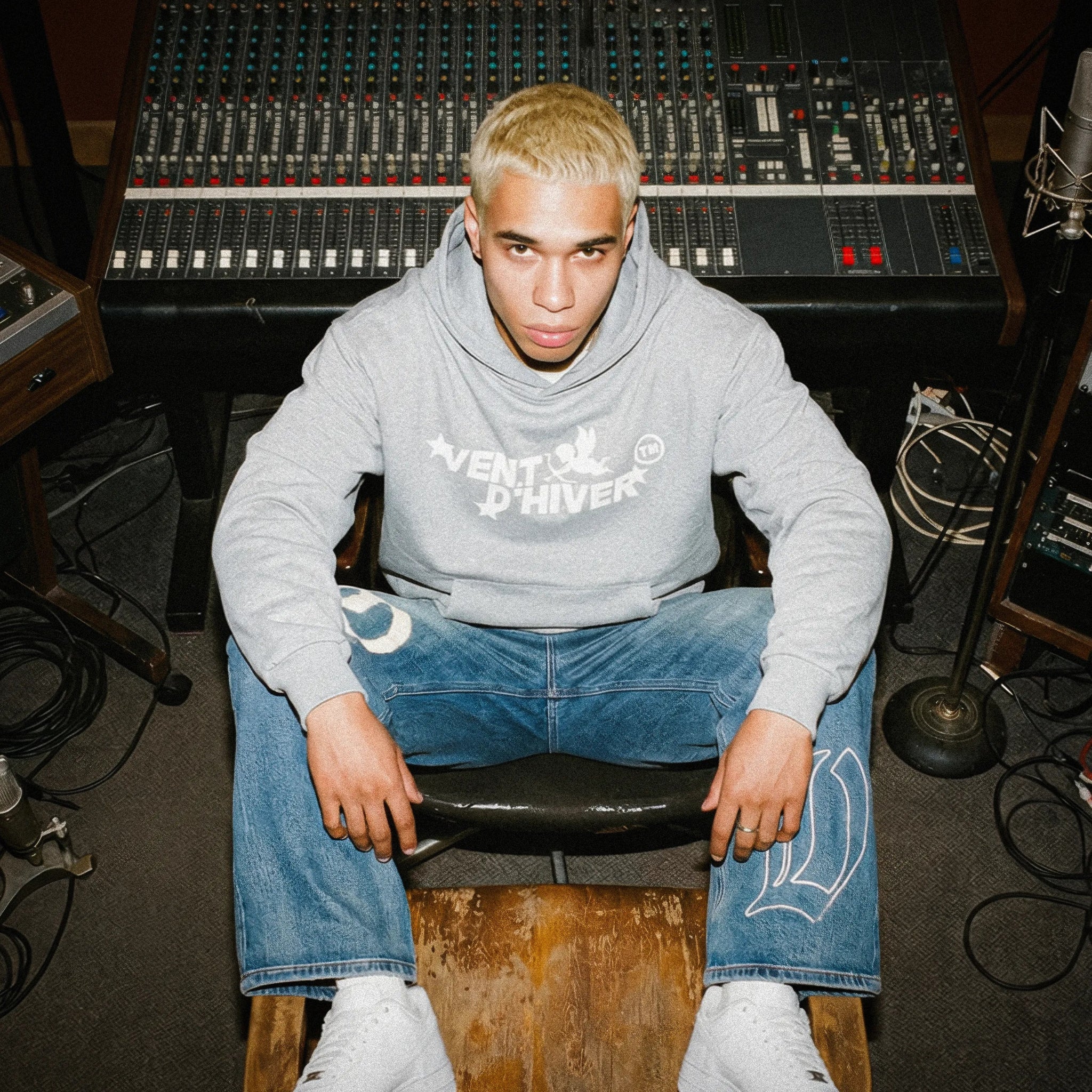 Person wearing a gray hoodie by VENT D'HIVER with text and blue jeans sitting in front of audio mixing equipment.