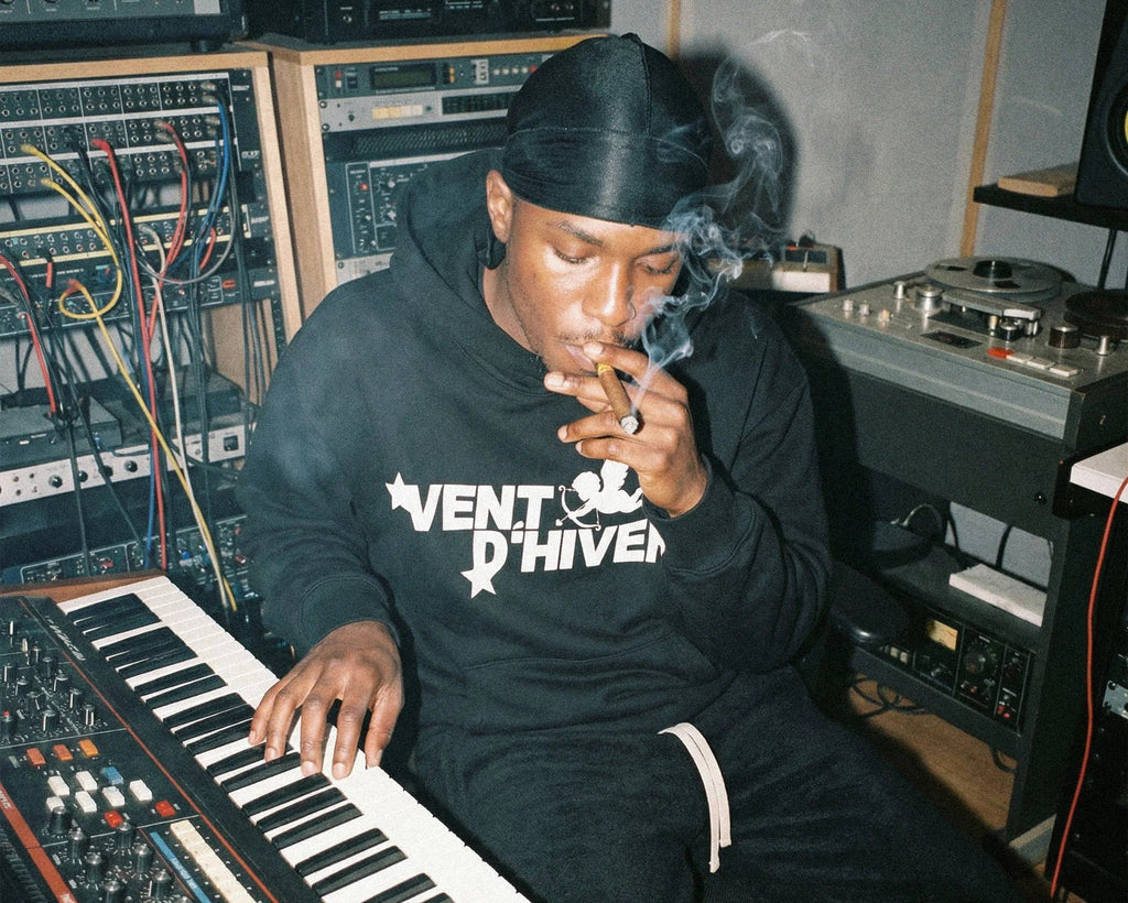 Person smoking a cigarette while sitting at a keyboard in a studio setting wearing hoodie and sweatpants by VENT D'HIVER