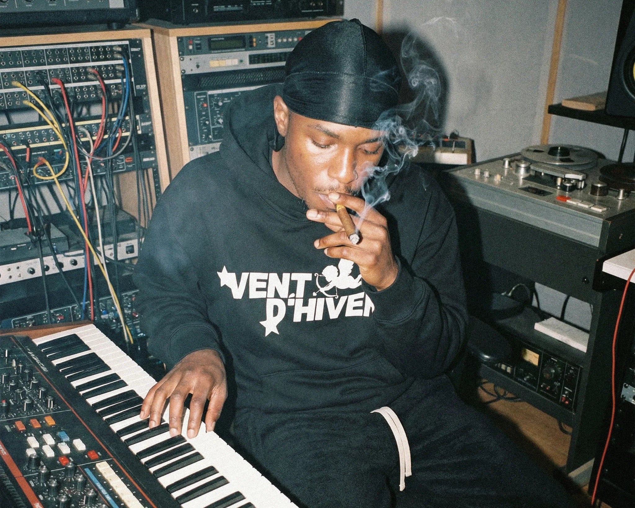 Person smoking a cigarette while sitting at a keyboard in a studio setting wearing hoodie and sweatpants by VENT D'HIVER