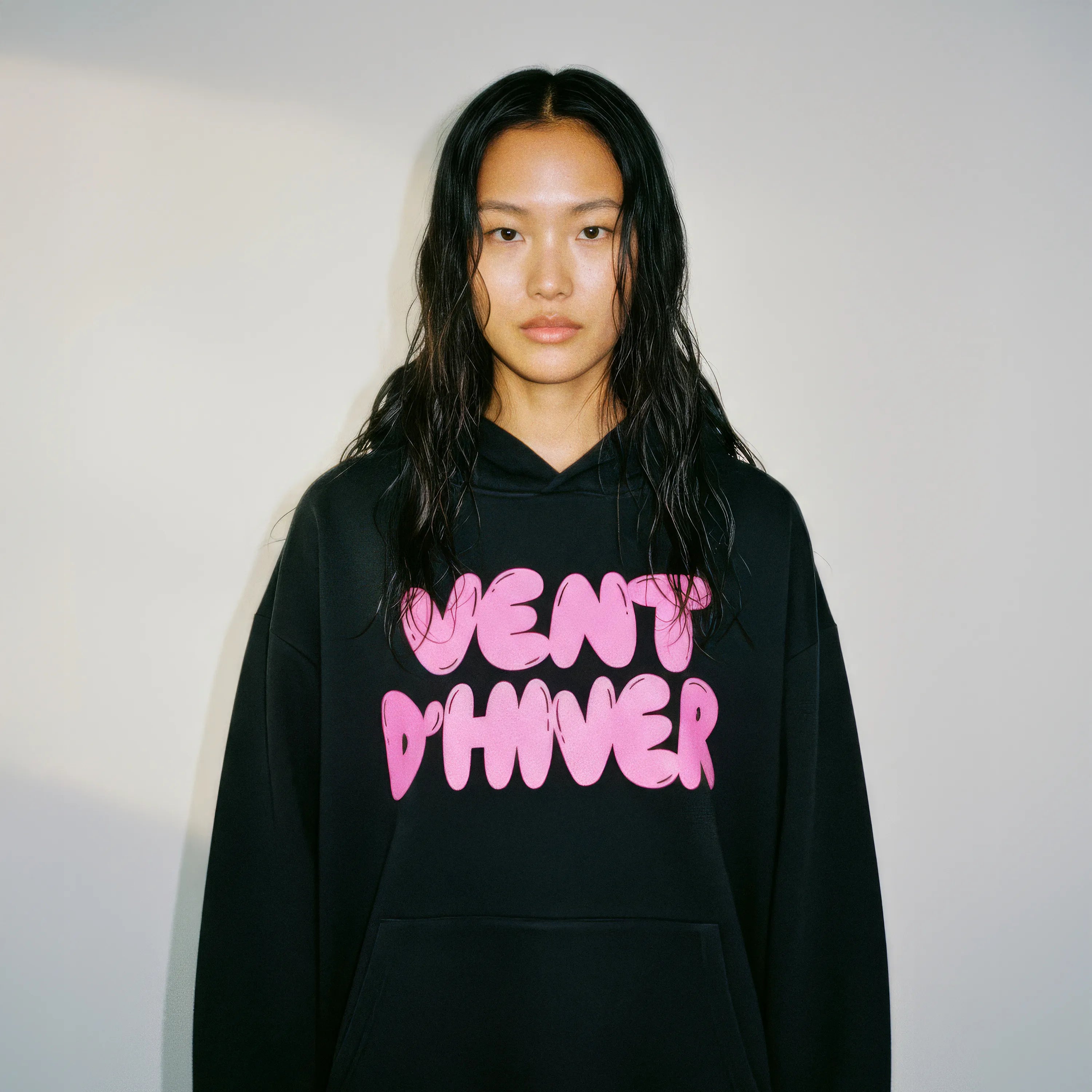 Person wearing a black hoodie by VENT D'HIVER with pink text against a plain background